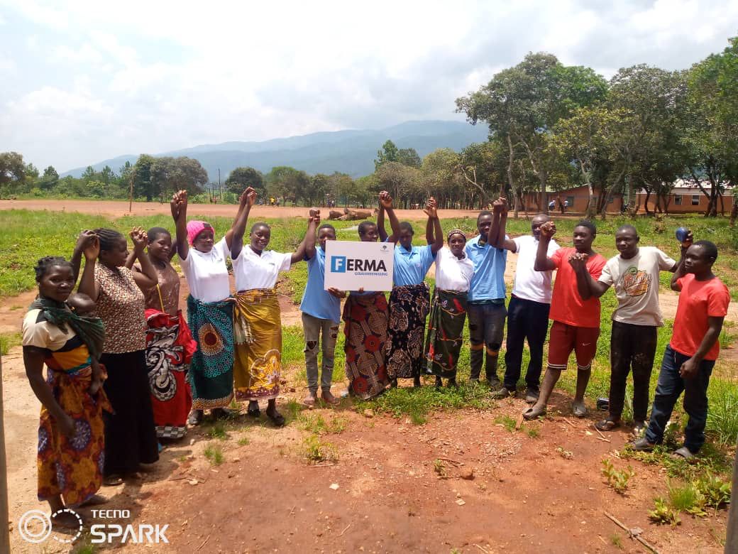 Eine Gruppe von Menschen in Malawi hält ein Schild mit dem Logo der FERMA Gebäudereinigung GmbH und jubelt. Im Hintergrund sind grüne Felder und Berge zu sehen.
