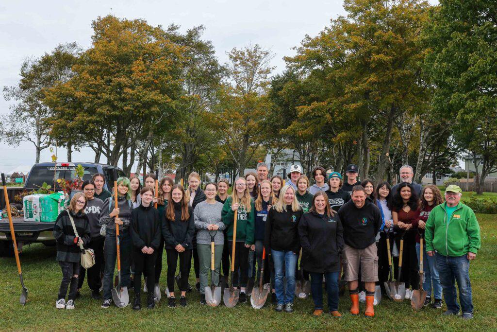 Gruppenfoto von Freiwilligen, die an einem Baumpflanzungsprojekt teilnehmen, ausgestattet mit Schaufeln und Gartengeräten, vor einem mit Pflanzen beladenen Pickup-Truck.