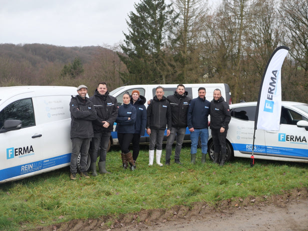 Eine Gruppe von acht Personen steht in einer Reihe vor mehreren Fahrzeugen der Ferma Gebäudereinigung auf einem grasbewachsenen Feld. Im Hintergrund sind Bäume zu sehen.
