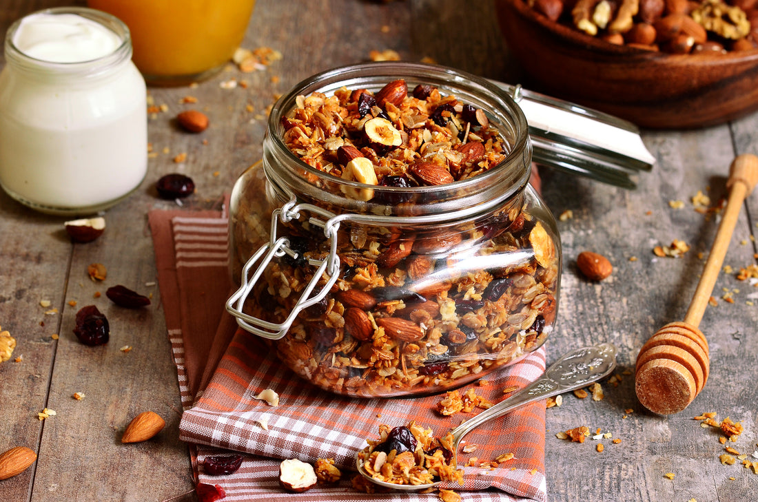 Das Bild zeigt ein Glas mit hausgemachtem Granola auf einem rustikalen Holztisch. Neben dem Glas liegen ein Löffel mit Granola, ein Honiglöffel und ein Glas Joghurt. Das Granola enthält Nüsse, getrocknete Früchte und Haferflocken.