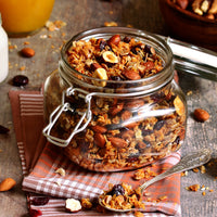 Das Bild zeigt ein Glas mit hausgemachtem Granola auf einem rustikalen Holztisch. Neben dem Glas liegen ein Löffel mit Granola, ein Honiglöffel und ein Glas Joghurt. Das Granola enthält Nüsse, getrocknete Früchte und Haferflocken.