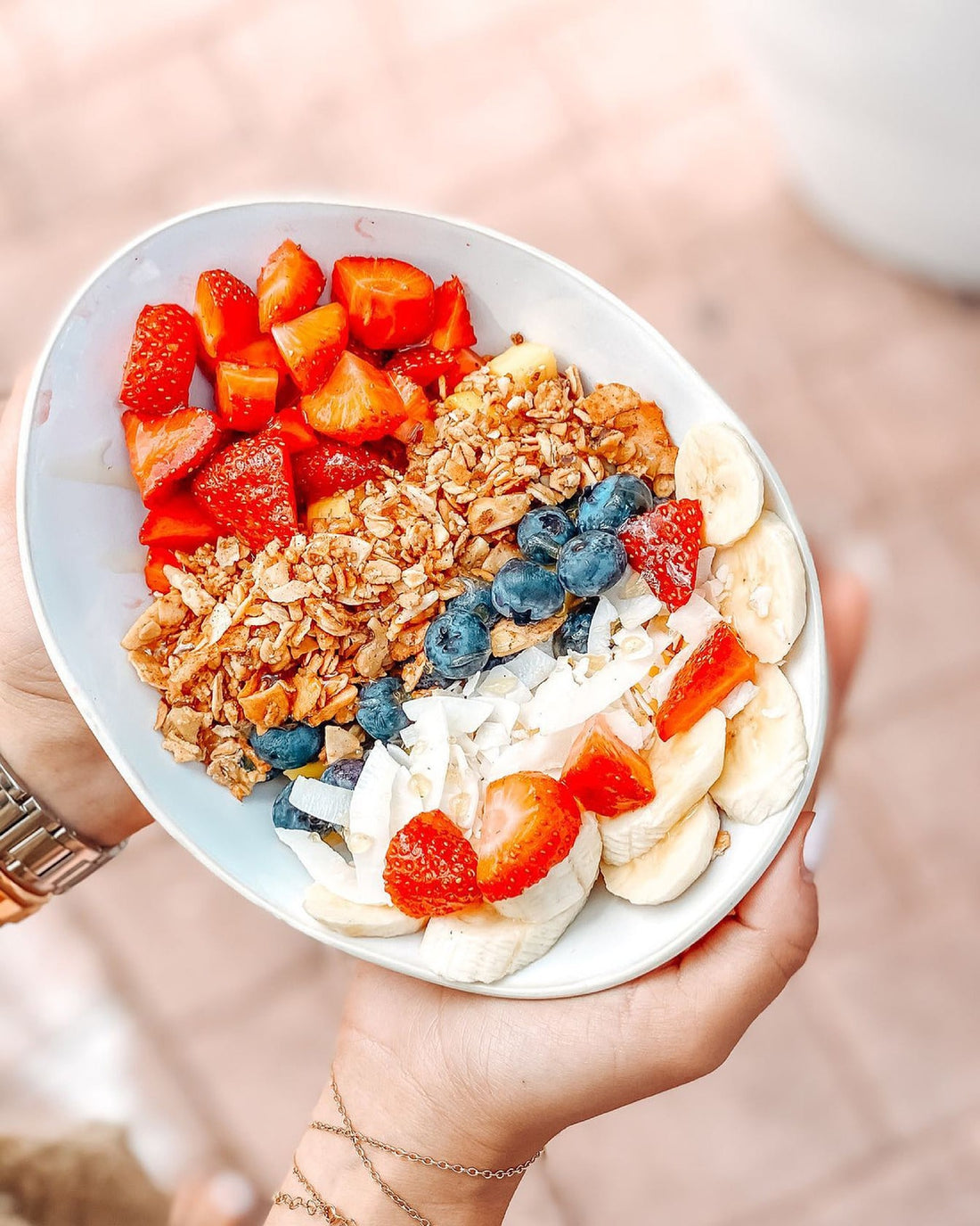 Das Bild zeigt eine Schüssel mit geschnittenen Erdbeeren, Bananenscheiben, Blaubeeren, Granola und Kokosflocken, die von einer Person in den Händen gehalten wird. Das Arrangement wirkt frisch und appetitlich.