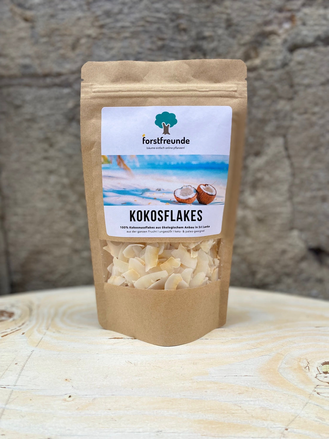 Eine Verpackung Forstfreunde Kokosflakes steht auf einem hellen Holztisch. Die braune, umweltfreundliche Verpackung zeigt ein Etikett mit dem Forstfreunde-Logo und einer Abbildung von Kokosnüssen vor einem Strandhintergrund. Die Kokosflakes stammen aus 100% ökologischem Anbau in Sri Lanka