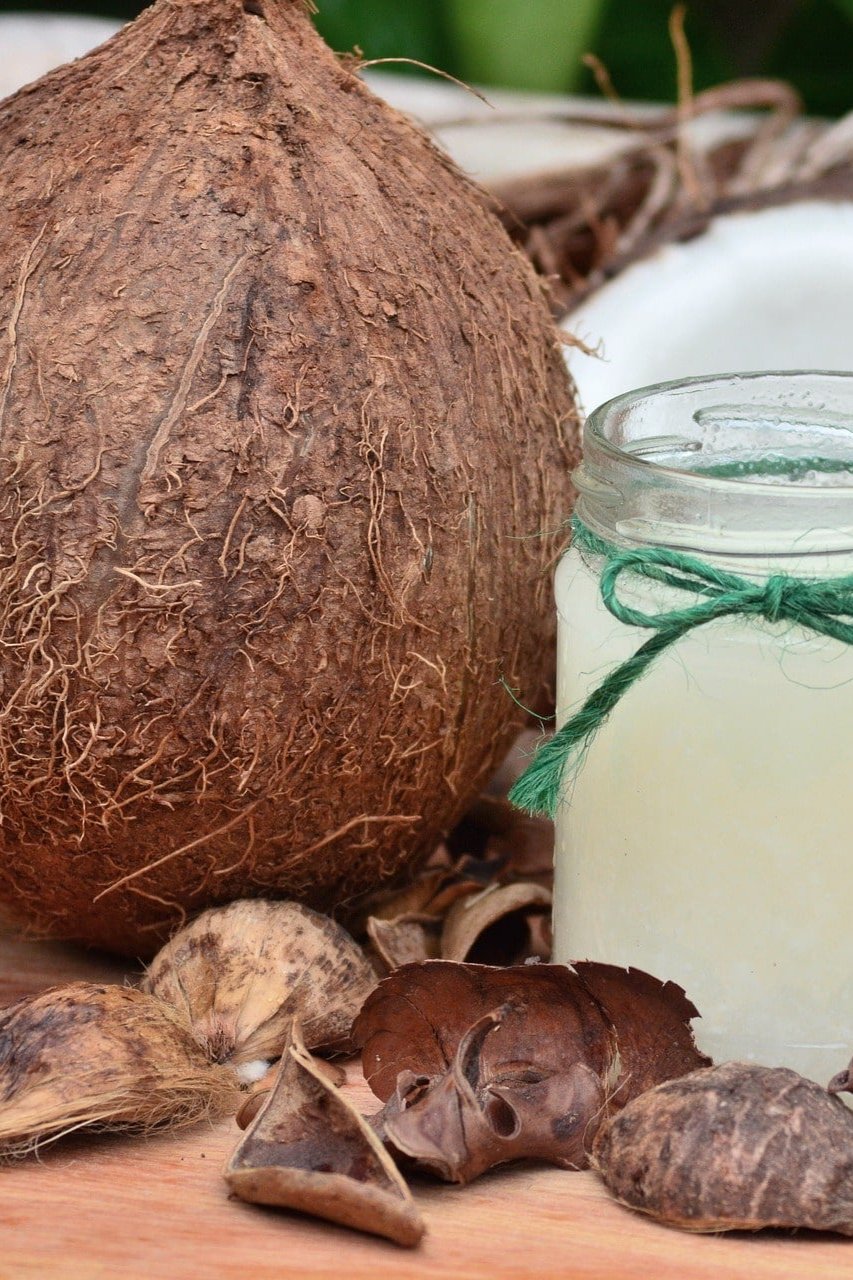 Das Bild zeigt eine ganze Kokosnuss, eine aufgebrochene Kokosnuss und ein Glas mit Kokosöl. Um die Kokosnüsse und das Glas herum liegen getrocknete Kokosnussstücke und -schalen. Im Hintergrund sind grüne Blätter unscharf zu erkennen.