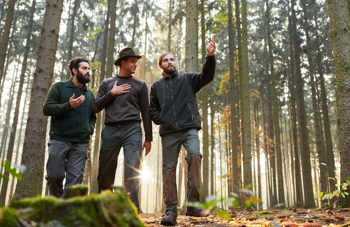 Drei Waldarbeiter diskutieren im Wald, einer zeigt mit der Hand auf etwas in der Ferne. Die Sonne scheint durch die Bäume im Hintergrund.