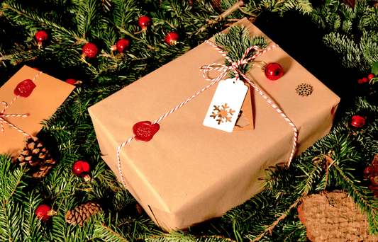 Zwei in braunes Papier eingepackte Weihnachtsgeschenke, verziert mit rot-weißen Schnüren, Wachssiegeln und Tannenzweigen. Die Geschenke sind auf einem Bett aus Tannenzweigen und roten Weihnachtsornamenten arrangiert.