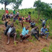 Eine Gruppe von Menschen sitzt auf einem Hügel, winkt und lächelt in die Kamera. Die Szene zeigt Freude und Gemeinschaftsgefühl bei einem Aufforstungsprojekt in Malawi.